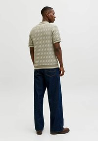 Ein Mann steht mit dem Rücken zur Kamera, trägt ein beiges, gemustertes Polohemd mit kurzen Ärmeln, dunkelblaue Jeans mit weitem Bein und braune Schuhe.