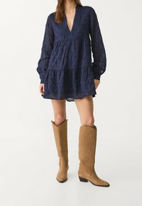 Robe en dentelle bleu marine avec des manches longues et un décolleté en V, présentant un design à volants, associée à des bottes de cowboy crème hauteur genou.
