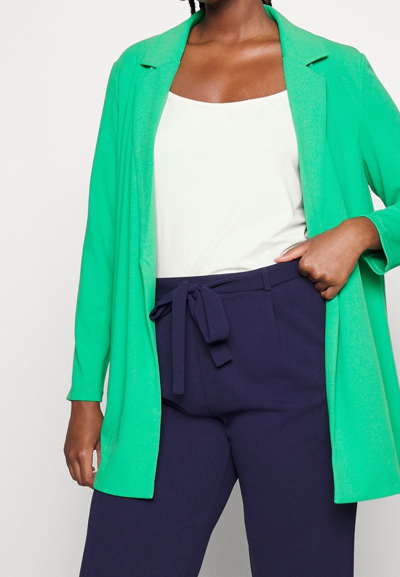 Zielony długi blezer noszony na białym topie na ramiączkach i ciemnogranatowych spodniach z wiązaniem z przodu. Blezer ma gładką fakturę i dopasowany krój.