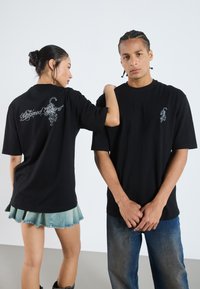 T-shirts en coton noir présentés sur deux modèles. Le dos arbore un design graphique blanc avec le texte "Esprit Indompté", et chacun a un petit logo à l'avant.