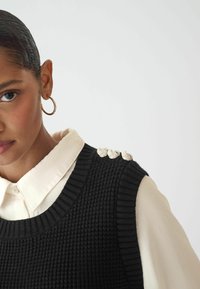Gilet en tricot noir avec un motif texturé, featuring trois accents décoratifs en argent sur l'épaule et une chemise blanche à col en dessous.