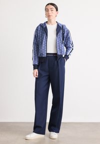 Giacca blu a motivi tagliata con pantaloni blu navy. La giacca ha un cappuccio e polsini a righe. Una t-shirt bianca e sneakers bianche completano l'outfit.