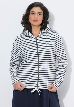 Kvinde med mørkt hår iført en hvid hættetrøje med sort horisontale striber og et navyblåt skørt, stående foran en ensfarvet hvid baggrund.