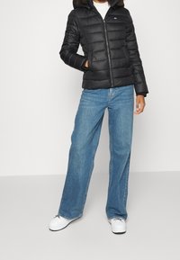 Czarna kurtka puchowa z kapturem wykończonym futrem, zapinana na zamek, z poziomym przeszyciem, noszona z szerokimi niebieskimi dżinsami i białymi trampkami.