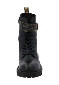 Botín de cuero negro con cordones en la parte frontal, una correa acolchada con acentos dorados y una suela de goma gruesa. Textura suave y puntera redondeada.