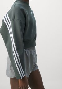 Mörkgrön sweatshirt med vita ränder på ärmarna, tillsammans med grå shorts med elastisk midja. Tyget verkar slätt och avslappnat.