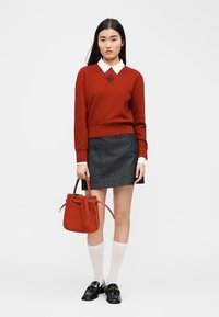 Femme portant un pull rouge sur une chemise blanche à col, une mini-jupe foncée, des chaussettes blanches montantes jusqu'aux genoux, des mocassins noirs, tenant un sac à main rouge.