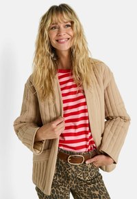 Femme souriante portant une veste matelassée beige, une chemise rayée rouge et rose, un pantalon à imprimé léopard et une ceinture marron avec une boucle en or.