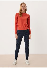 Czerwony bawełniany sweter z okrągłym dekoltem, zestawiony z granatowymi spodniami o slim fit. Model nosi białe sneakersy, stojąc na neutralnym tle.