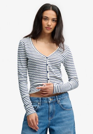 Jeune femme portant un haut à manches longues blanc et bleu marine rayé, boutonné, et un jean bleu taille haute, posant en ajustant le haut d'une main.