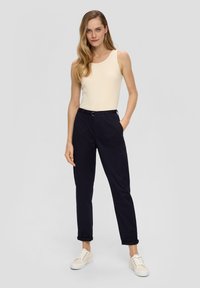 Débardeur beige à côtes, pantalon tapered bleu marine avec ourlets roulés et sneakers clairs. La tenue présente un design simple et une silhouette ajustée.