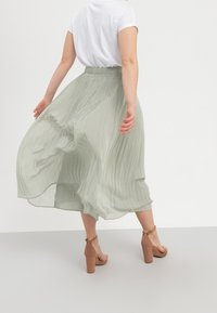 Gonna midi plissettata in tessuto verde chiaro, caratterizzata da una silhouette fluida e una vita elastica. Indossata con una maglietta bianca e sandali con tacco color cuoio.