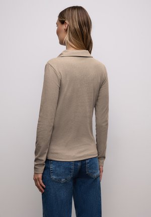 Mujer con una camisa de manga larga beige y jeans azules, de pie de espaldas contra un fondo claro y liso.