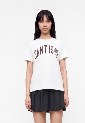 Jeune femme aux longs cheveux foncés portant un t-shirt blanc avec le texte "GANT 1949" et une jupe plissée noire, debout devant un fond uni.