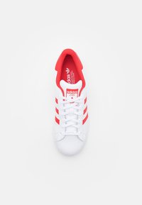 Zapatilla Adidas blanca con acentos rojos, tres rayas en el lateral y una punta texturizada. Presenta una lengüeta y una plantilla roja acolchadas.