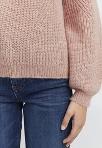 Maglione lavorato a maglia di un delicato rosa chiaro con una finitura texture e polsini a coste, abbinato a jeans in denim blu. Il maglione ha una vestibilità ampia.