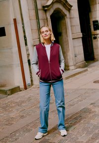 Maroon zip-up bomber jacket with gray sleeves, ribbed cuffs, white t-shirt underneath, paired with light blue jeans and white sneakers.