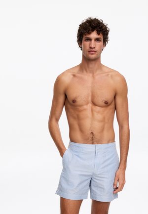 Młody mężczyzna z kręconymi włosami stoi bez koszulki, nosząc jasnoniebieskie i białe spodenki w paski na jednolitym białym tle.