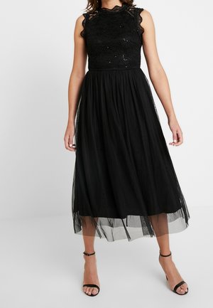 Femme portant un haut en dentelle noire sans manches et une jupe midi fluide en tulle avec des sandales à talons noirs à bride à la cheville.