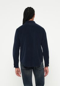 Camisa de pana de color azul marino con mangas largas, puños abotonados y dobladillo recto. Textura suave con costuras y detalles de diseño mínimos.