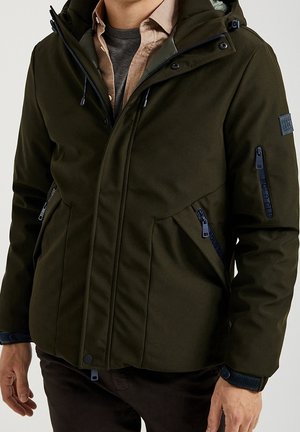 Homme portant une veste matelassée vert foncé avec poches zippées et capuche, superposée à un t-shirt gris et une chemise beige.