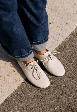 Piedi con scarpe bianche con lacci e jeans blu scuro risvoltati che stanno su un pavimento testurizzato con sezioni bianche e marroni.