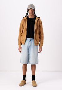 Haut à capuche zippé marron, t-shirt noir, shorts en denim clair, chaussettes noires et baskets beiges. Casquette grise avec un logo. Tenue simple et décontractée.