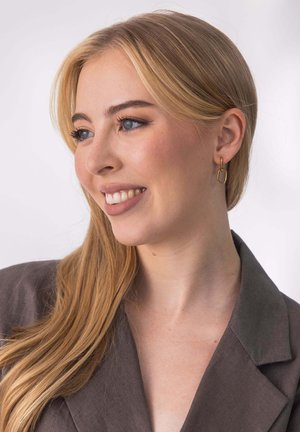 Goldene geometrische Ohrringe mit rechteckigem Design. Das Modell hat lange blonde Haare und trägt einen dunkelgrauen Blazer. Heller Hintergrund.