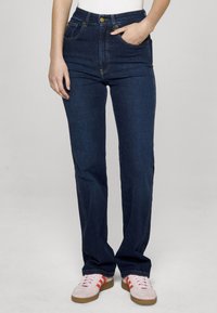 Temno modre jeans hlače iz jeansa z visokim pasom, ravnimi hlačnicami, petimi žepi in zlatimi kovinskimi dodatki. Nošene z rožnatimi supergami.