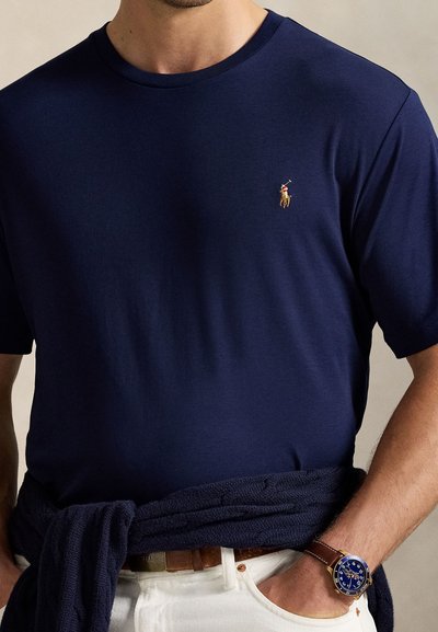 Bărbat purtând un tricou bleumarin cu guler rotund cu un mic jucător de polo brodat, pantaloni albi, ceas de piele maro și un pulover bleumarin legat în jurul taliei.