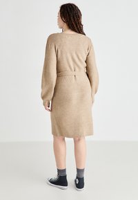 Vestido de punto beige con mangas largas y cintura ceñida. Presenta una textura acanalada y un corte recto que cae justo por encima de la rodilla.