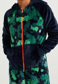 Monos de forro polar azul marino y verde con patrones abstractos, cierre de cremallera, cremallera naranja y bolsillos frontales. Cuenta con puños de canalé y capucha.