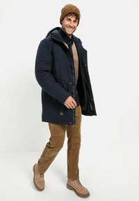 Marineblaue Steppjacke mit Kapuze, getragen über einem hellbraunen Pullover und braunen Cordhosen. Hellbraune Stiefel mit robuster Sohle, lächelndes Model.