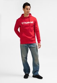 Felpa rossa con cappuccio e logo bianco, materiale in cotone, maniche lunghe, tasca a marsupio. Abbinata a jeans di denim blu e scarpe da ginnastica nere, in piedi su uno sfondo uniforme.