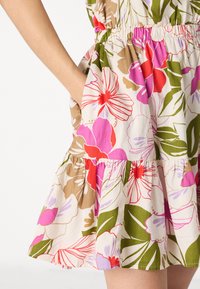 Blumenkleid mit einem crèmefarbenen Hintergrund, verziert mit lebhaften rosa, roten und grünen Blumen, geraffter Taille und Rüsche am Saum. Seitenfach sichtbar.