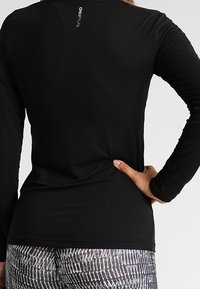 Person mit einem figurbetonten schwarzen Langarmoberteil mit dem "ONLY PLAY" Logo auf dem oberen Rücken und gemusterten schwarz-weißen Leggings, die Hand auf der Hüfte.