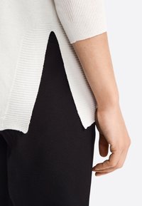 Top en tricot blanc à côtes avec des manches courtes, présentant un design à fente sur le côté, associé à un pantalon noir ajusté. Texture douce avec une silhouette épurée.