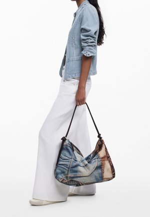 Femme portant un blazer à rayures bleu clair et un pantalon large blanc, tenant un grand sac à bandoulière en patchwork de denim et cuir marron.