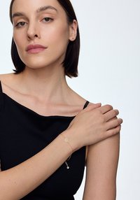 Złota bransoletka o filigranowym wzorze, z wiszącym charmem. Noszona na nagim ramieniu, na tle czarnej bluzki i neutralnego tła.