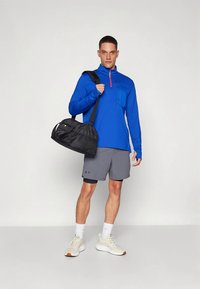 Bărbat purtând bluză albastră cu mâneci lungi, pantaloni scurți gri, adidași albi, cu o geantă neagră de sală peste umăr, stând în picioare pe un fundal simplu.