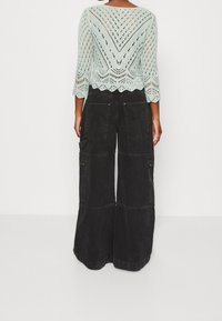 Rückansicht einer Person, die ein hellgrünes gestricktes Oberteil mit 3/4-Ärmeln und schwarze weit geschnittene Hose mit großen Taschen und Schlaufen trägt.