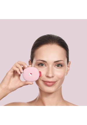 Femme aux cheveux tirés en arrière tenant un compact Hello Kitty rose près de son visage sur un fond rose clair.