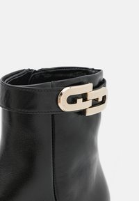 Bottine noire en cuir avec un détail de boucle rectangulaire dorée sur une bride près du bord supérieur sur fond blanc.