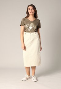 T-shirt vert olive avec un graphisme de cœur métallique, jupe midi crème avec un motif texturé, et baskets blanches avec accents rouges.