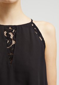 Haut noir sans manches avec une empiècement en dentelle au niveau de l'encolure et des épaules, avec une légère ouverture en forme de V à l'avant. Texture de tissu lisse.