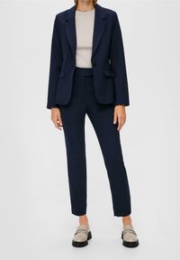 Námořnický modrý blazer s vroubkovanými klopami a dvěma předními kapsami, spárovaný s odpovídajícími kalhotami. Světle béžový top a lesklé béžové mokasíny.