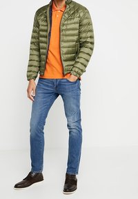 Giacca puffa verde oliva con colletto alto, polo arancione, jeans blu e scarpe marrone scuro, che mostrano una texture trapuntata e un design aderente.