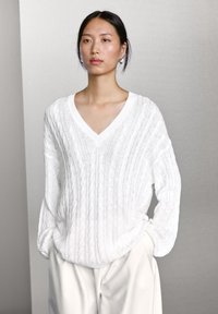 White knitted sweater with a V-neck, ribbed texture, and loose fit. Features cable knit design, long sleeves, and subtle droplet accent at hem.