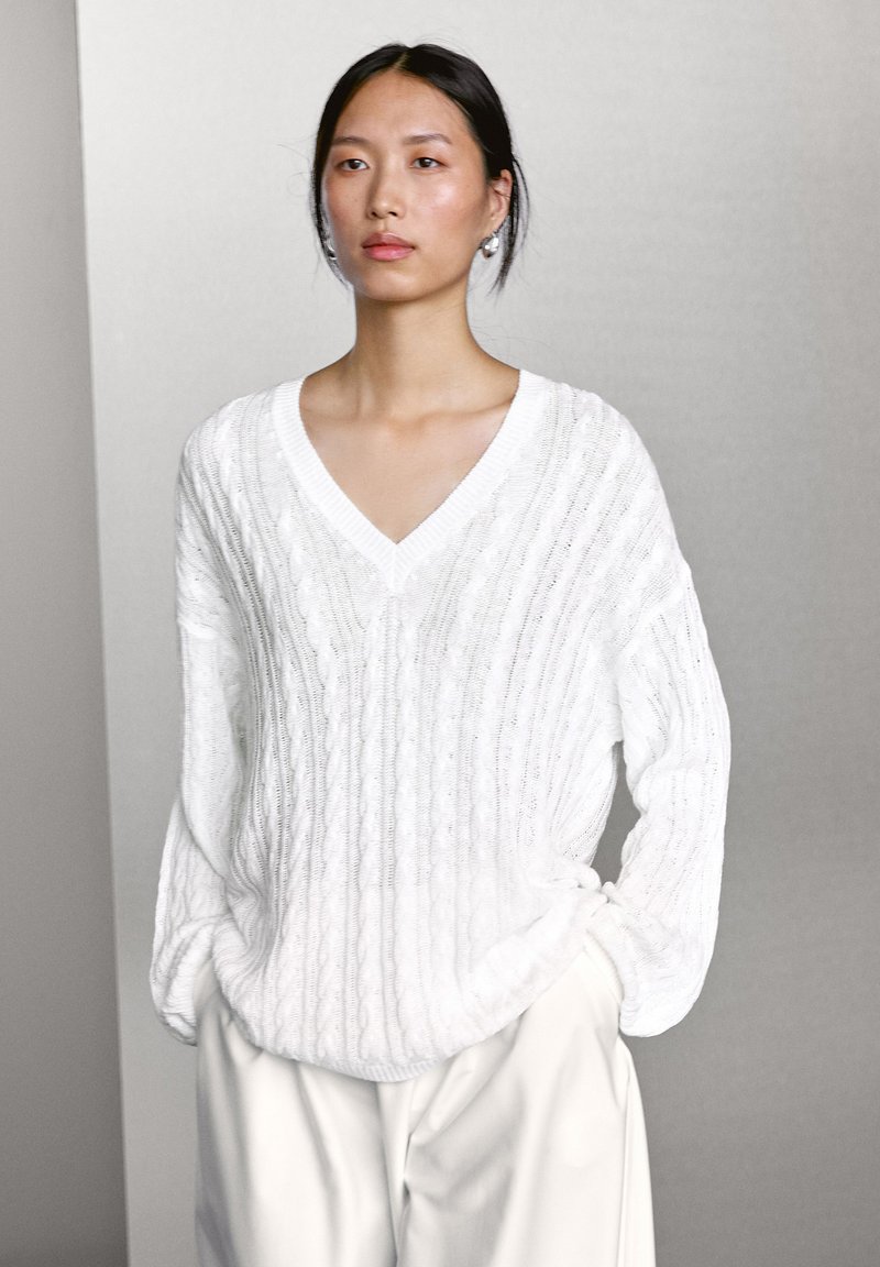 White knitted sweater with a V-neck, ribbed texture, and loose fit. Features cable knit design, long sleeves, and subtle droplet accent at hem.
