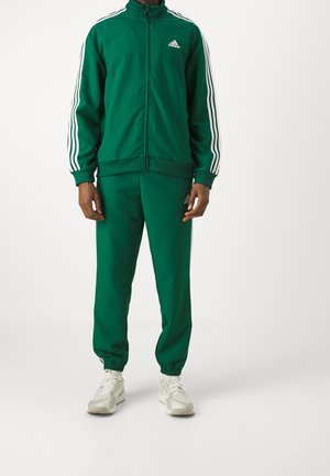 Homme portant un survêtement Adidas vert foncé avec des rayures blanches sur les manches et les jambes, associé à des baskets blanches, debout devant un fond uni.